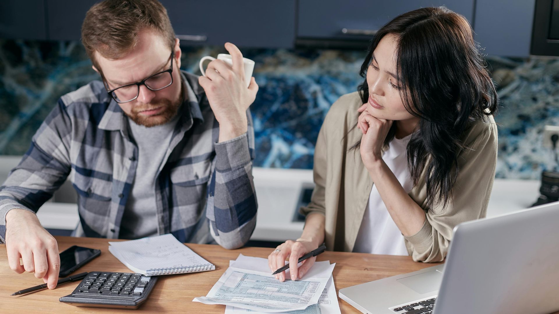 A couple reviews bills and documents on a laptop while discussing household finances.