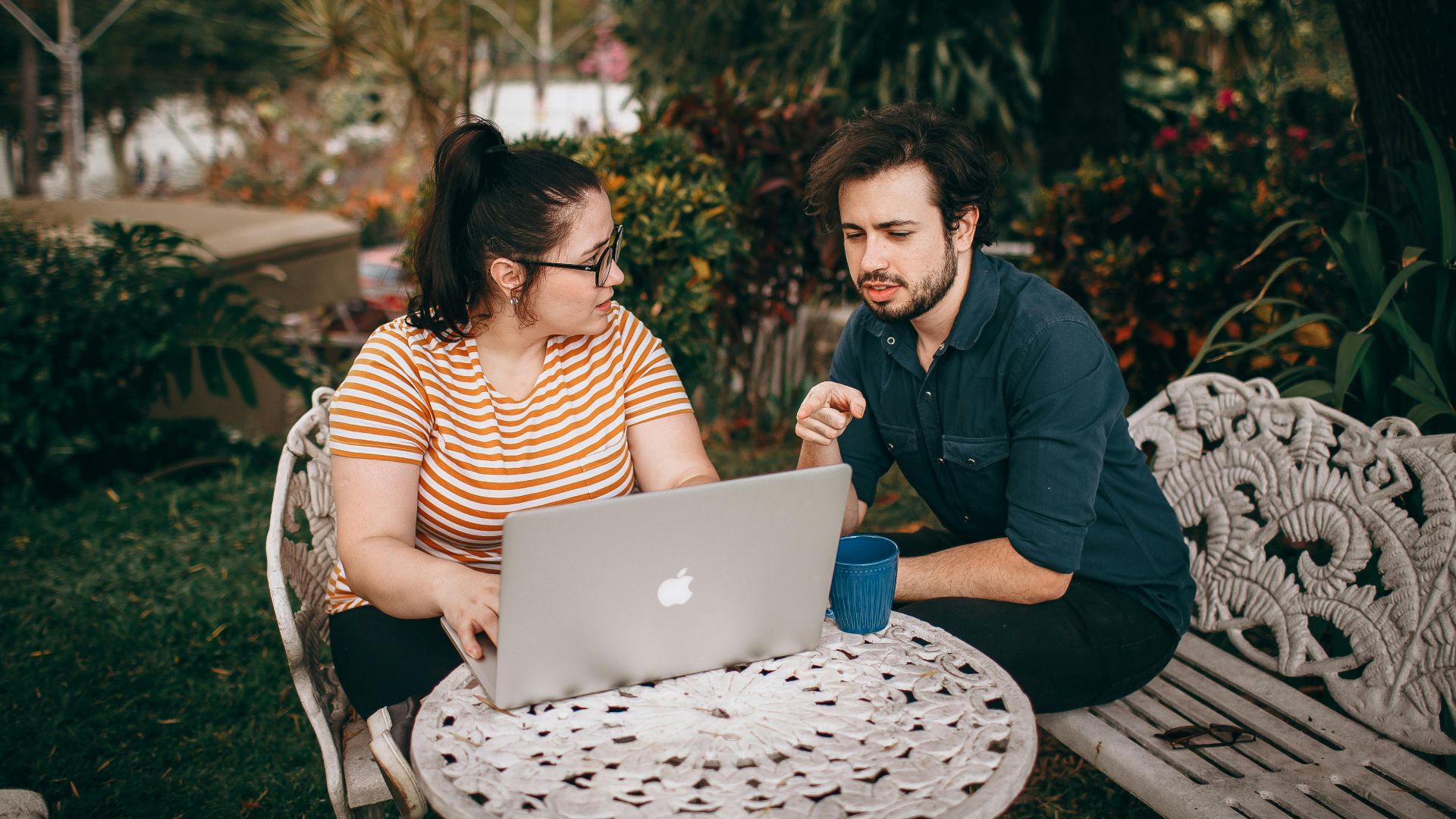 A man and woman working together on a laptop outdoors in a lush garden.