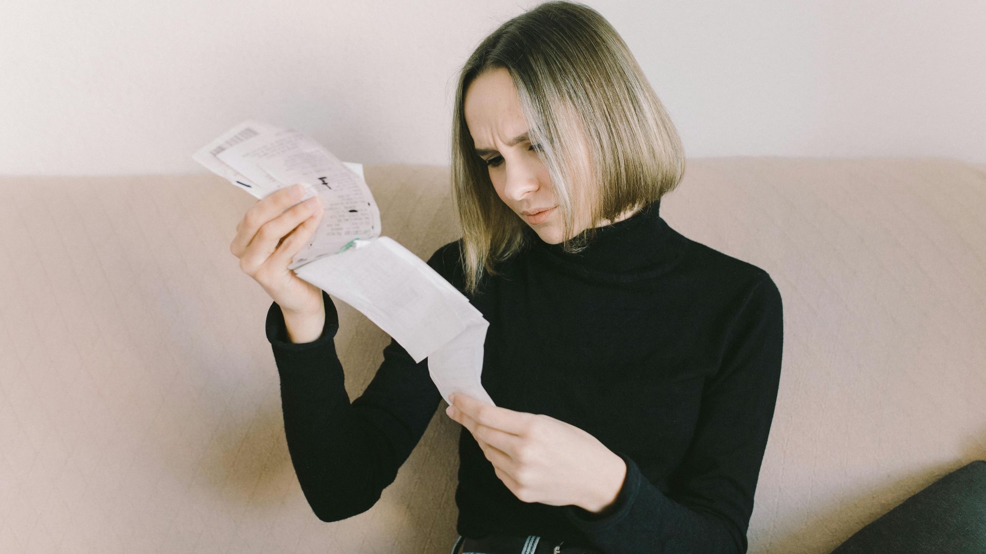 Concerned woman in black sweater examining bills on beige sofa indoors.