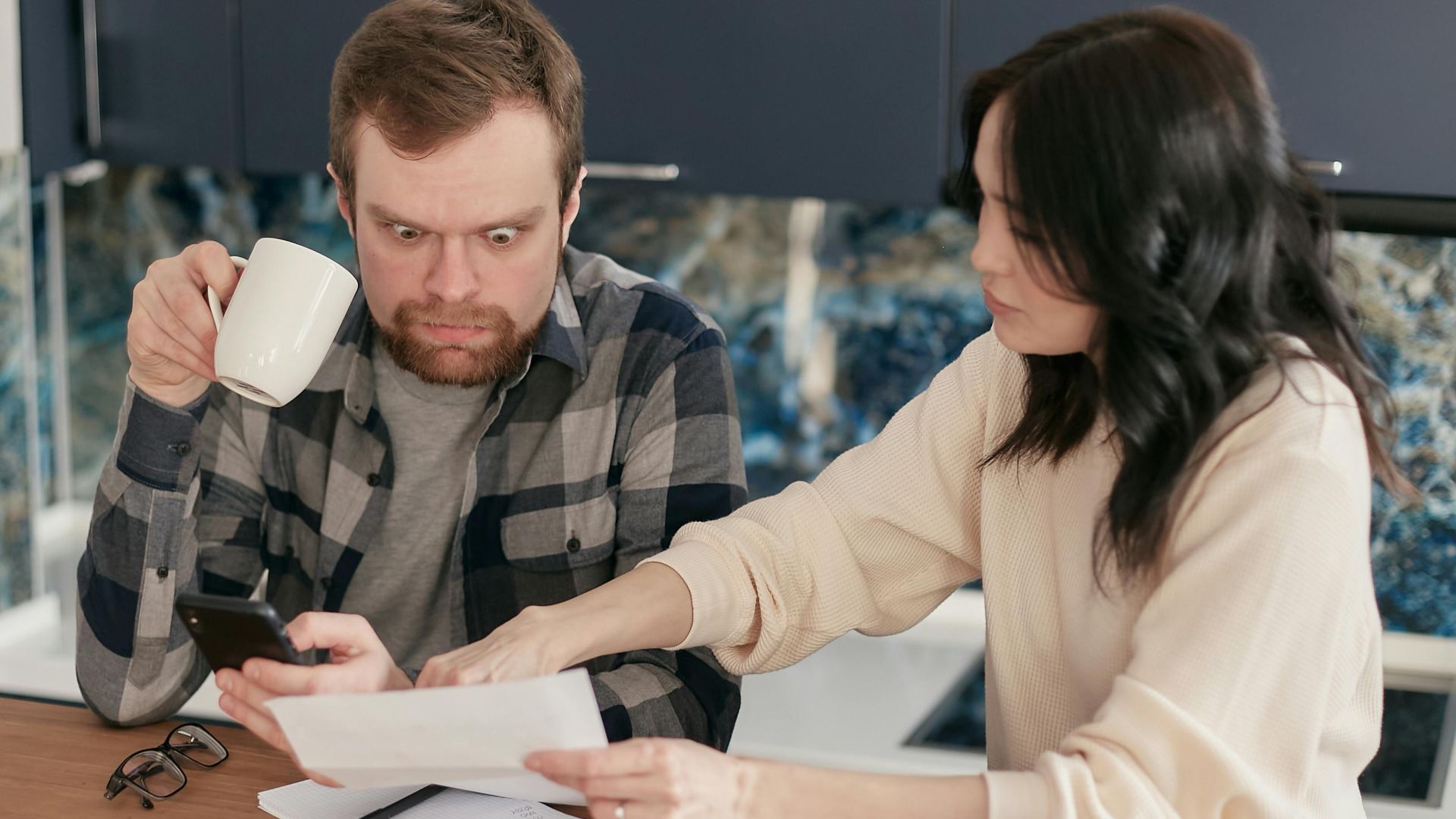 A couple reviews household bills in a kitchen, appearing concerned and focused.