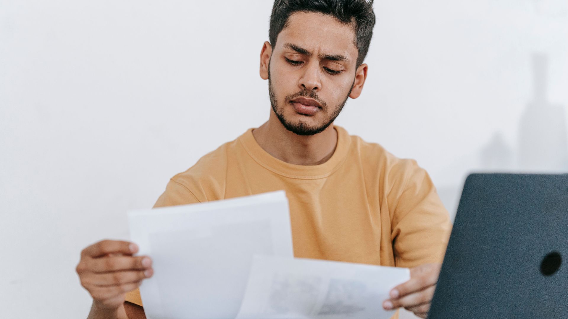 Attentive young bearded Hispanic male remote worker examining papers with results of project while working with laptop in home office