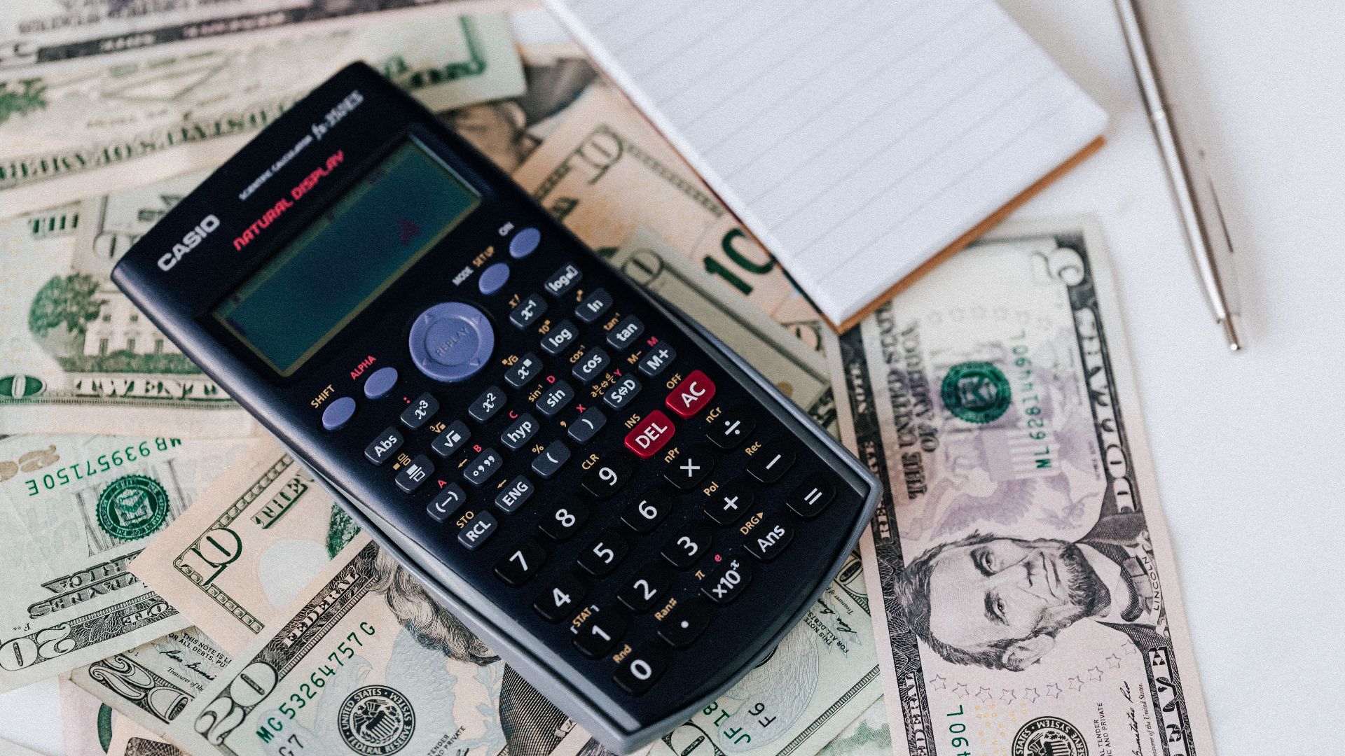 From above electronic calculator and notepad placed over United States dollar bills together with metallic pen for budget planning and calculation