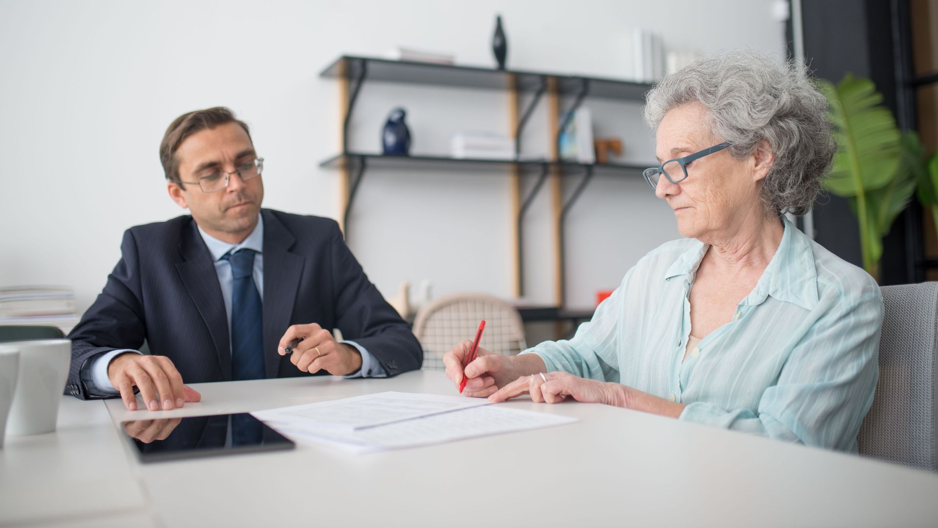Elderly woman signing paperwork in modern office with consultant.