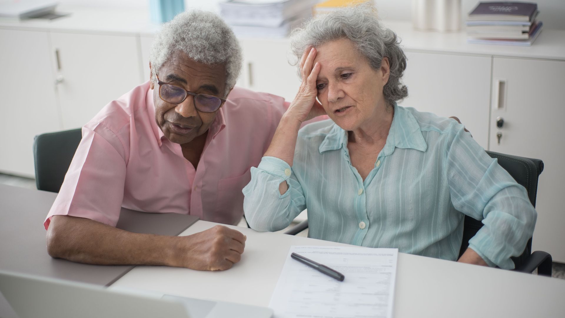 Elderly couple engaged with a laptop, reviewing paperwork at home office desk.