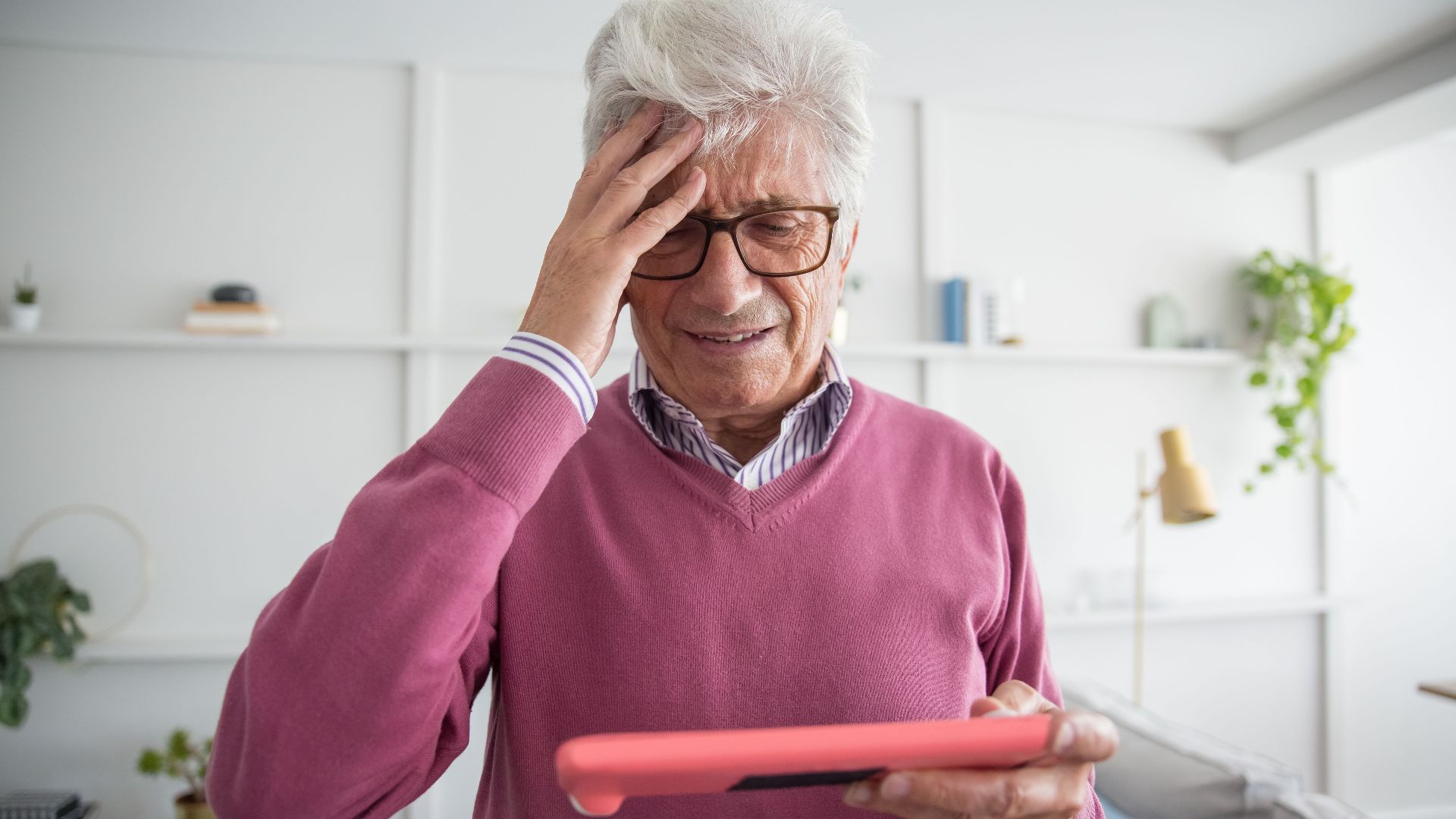 Elderly man in glasses using a tablet, puzzled expression, indoors.