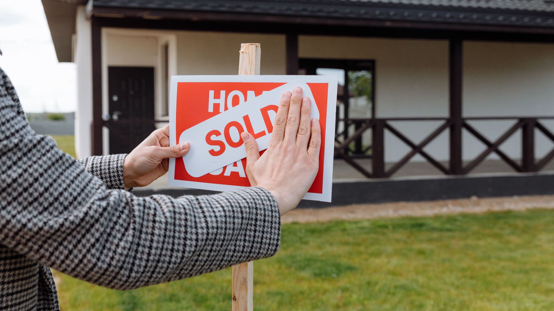 Close-up of hands placing 'sold' sticker on sign in front of a new house.