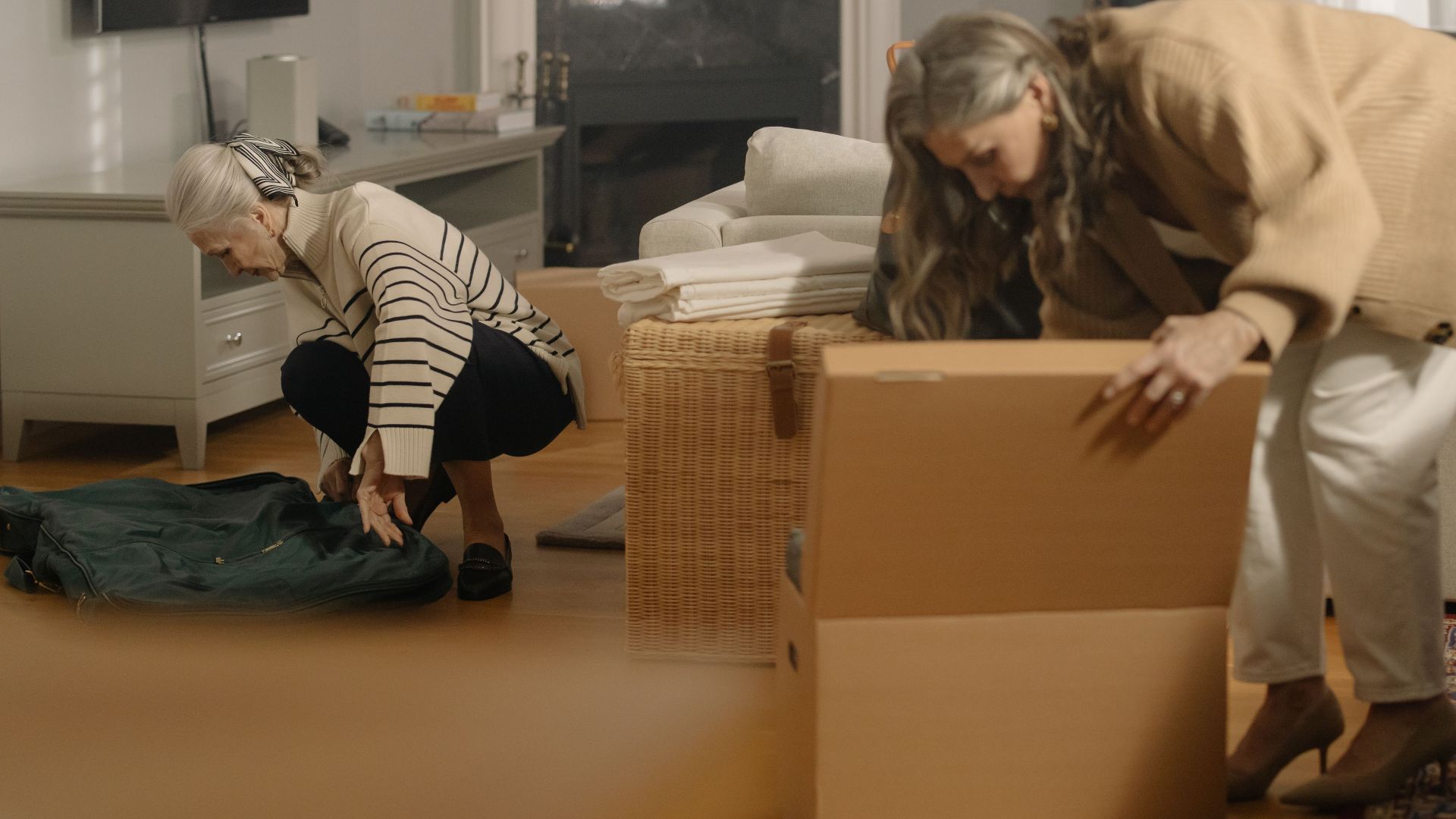 Two senior women packing belongings in boxes, preparing for a move in a cozy living room.