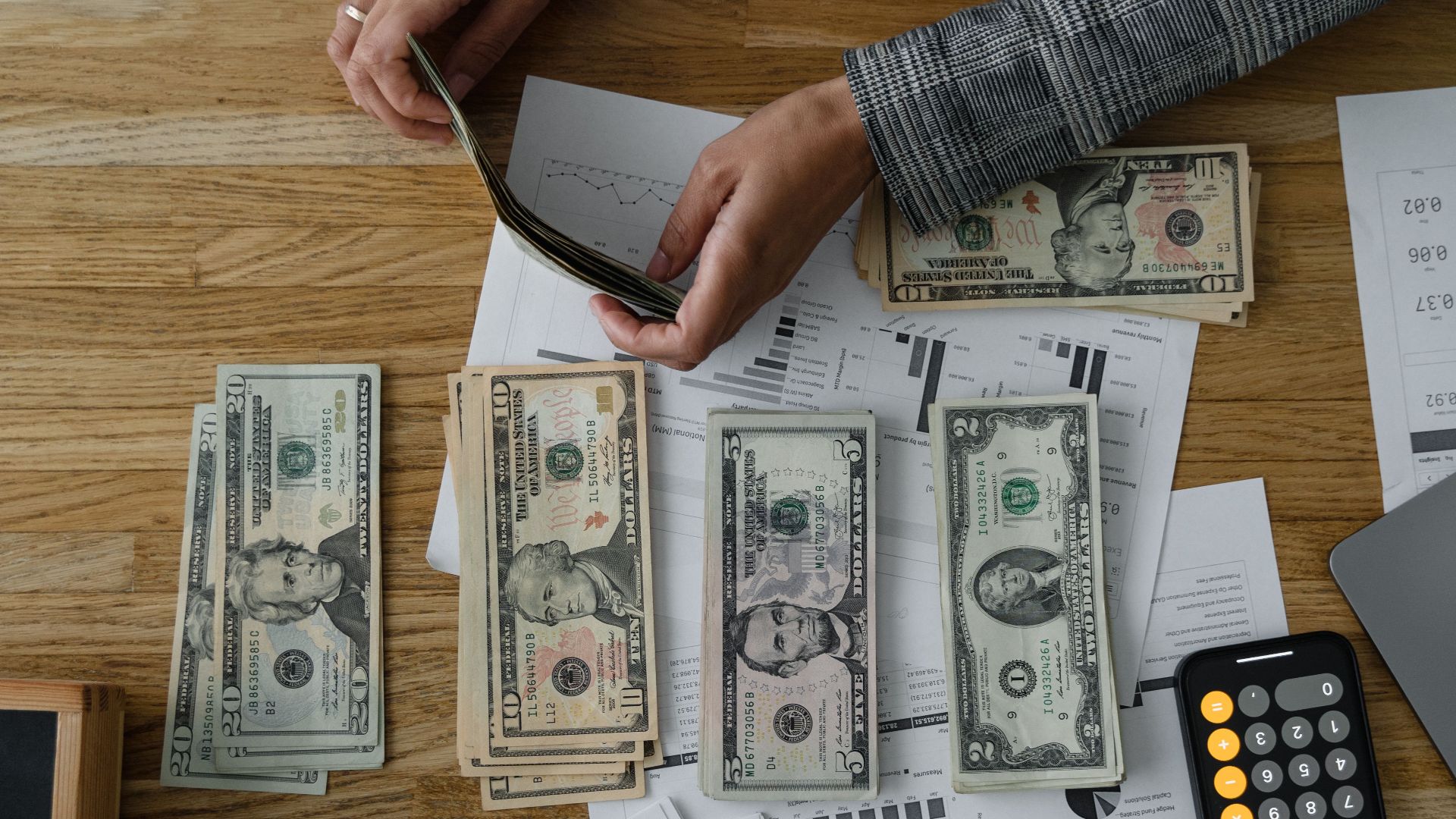 Person counting cash with a calculator and documents on a wooden desk, representing financial planning.