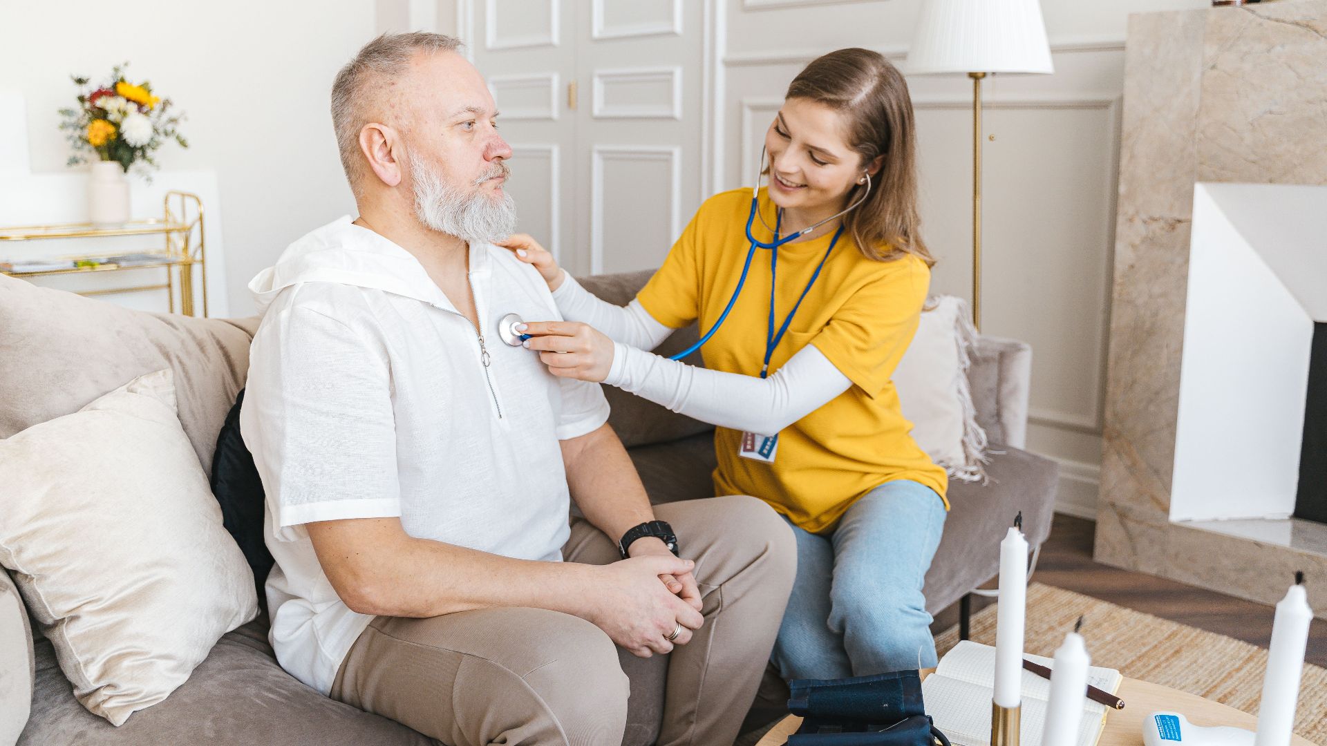 A nurse uses a stethoscope for a home check-up on a senior adult in a cozy living room.