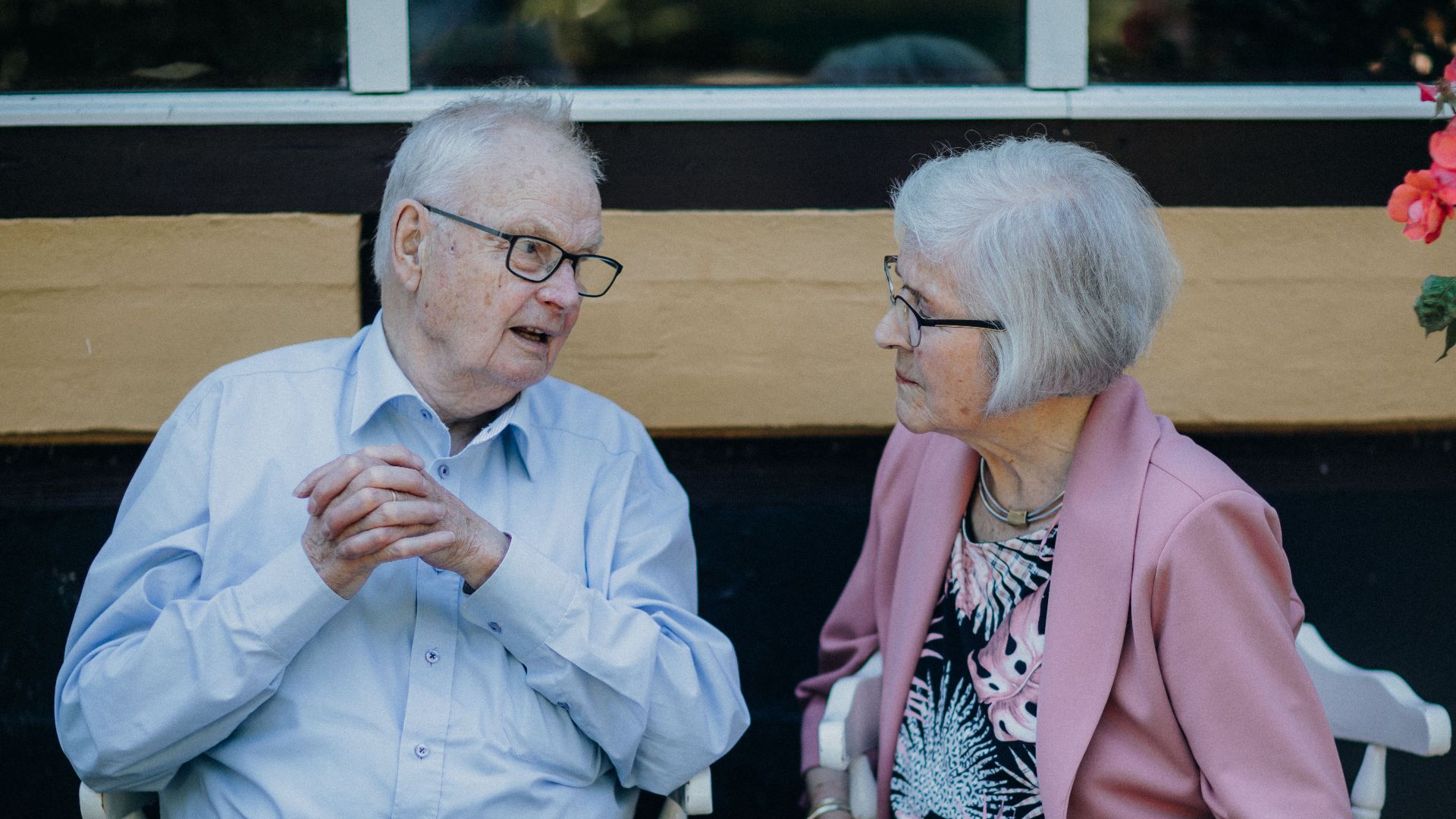 Senior couple sitting outside, engaged in conversation with flowers nearby. Captures a candid moment.