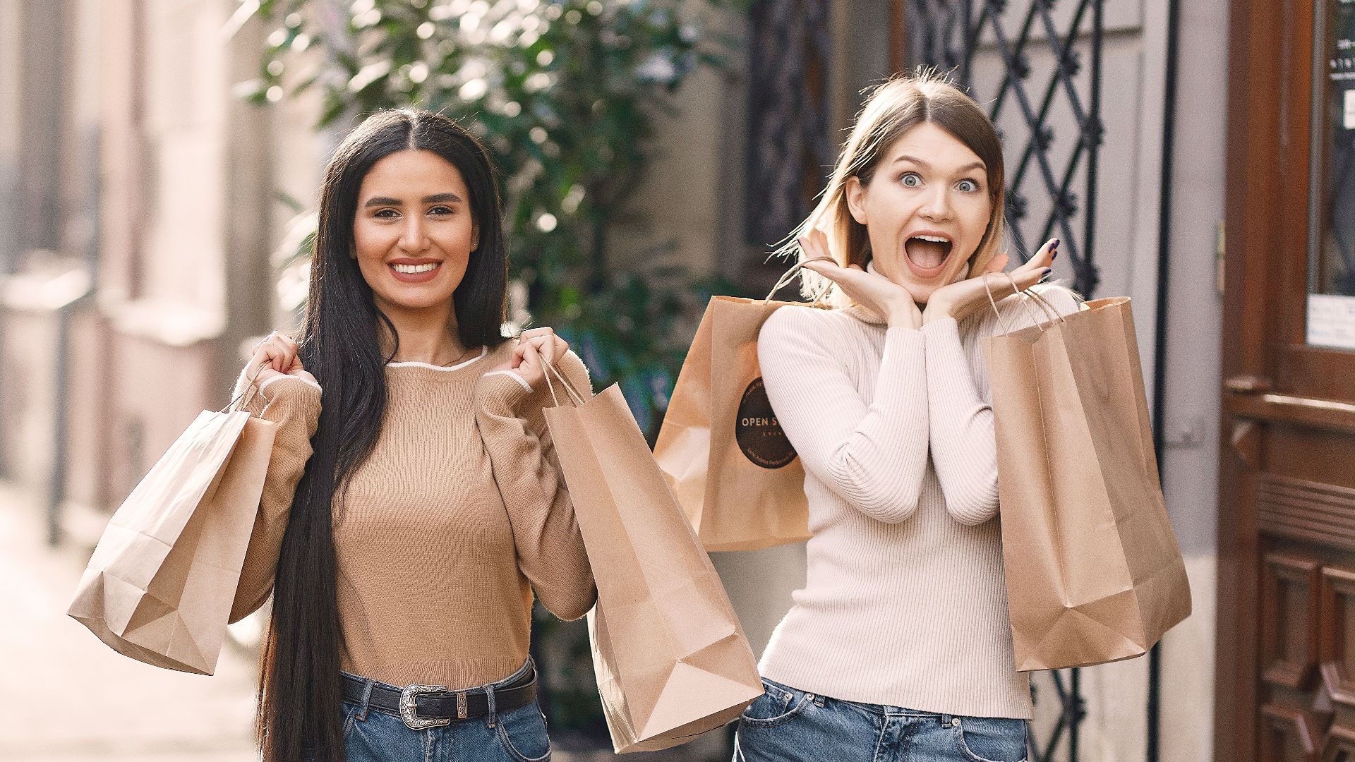 Positive young casually dressed women with shopping bags standing near building while smiling and looking at camera in sunny summer day