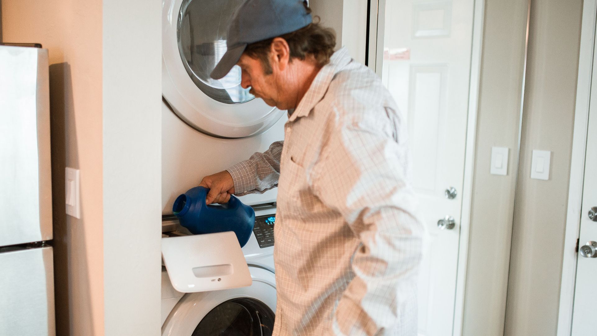 Man pouring liquid detergent into a stacked washer and dryer in a modern laundry room.