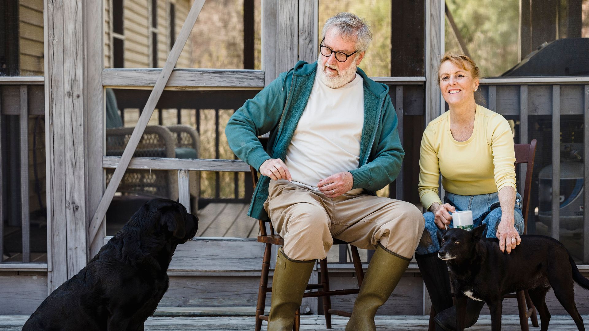 Delightful senior couple in rain boots sitting on chairs with black dogs while spending time together on terrace