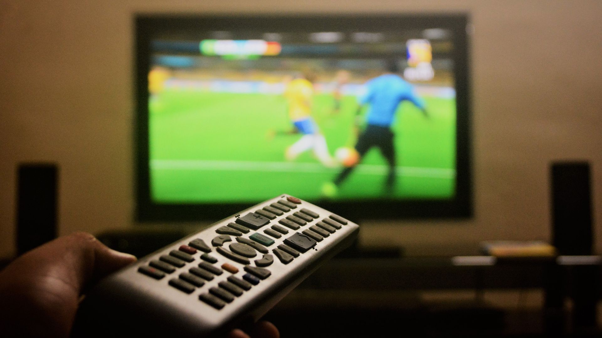 Close-up of a hand holding a remote control in front of a TV showing a football match.