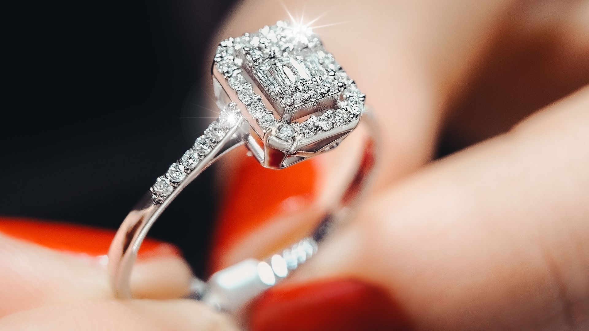 Close-up of a sparkling diamond engagement ring held by a person with red nails.