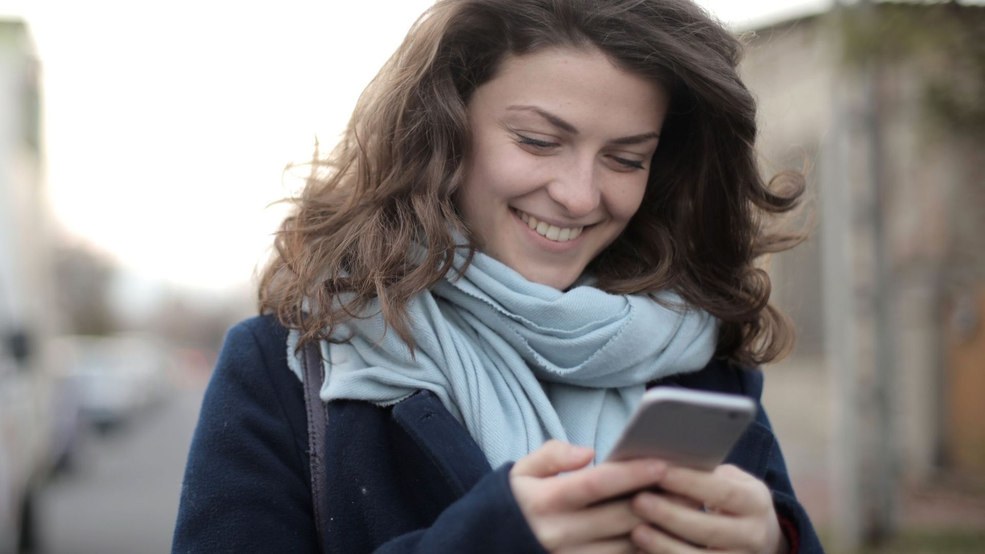 A young woman smiling while texting on her smartphone outdoors, wearing a scarf.