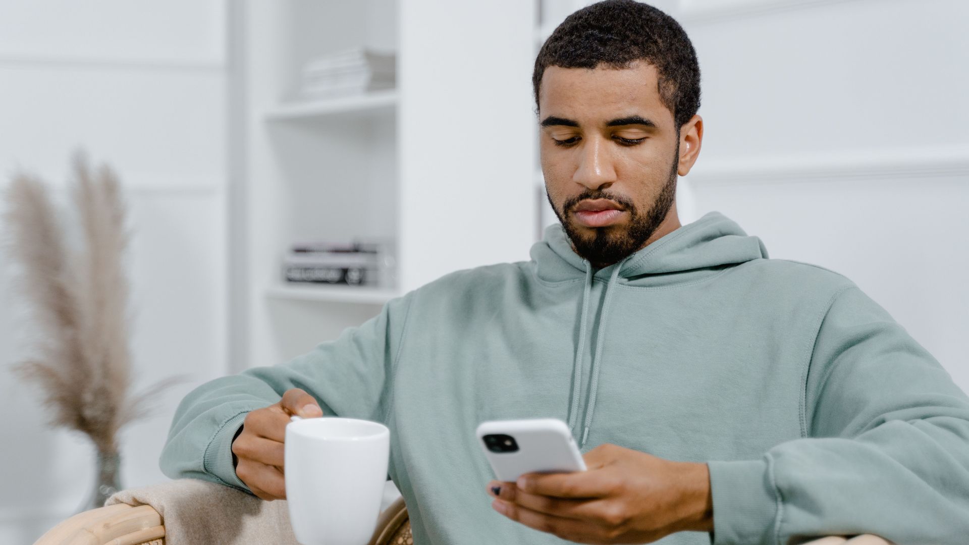 Man in a cozy setting using smartphone and holding a coffee mug.