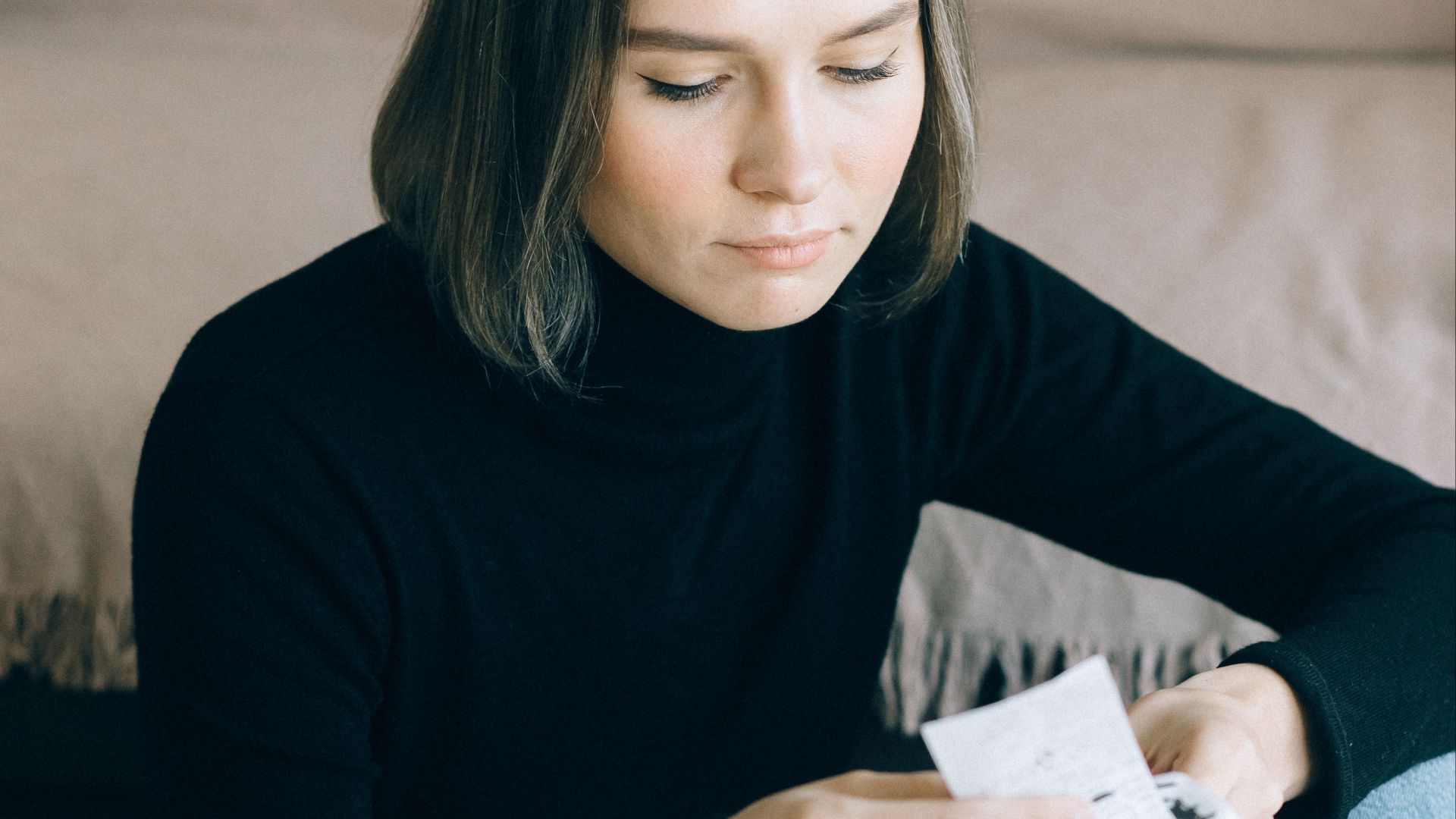 Young woman in black sweater reviewing bills at home, using a calculator, surrounded by documents.