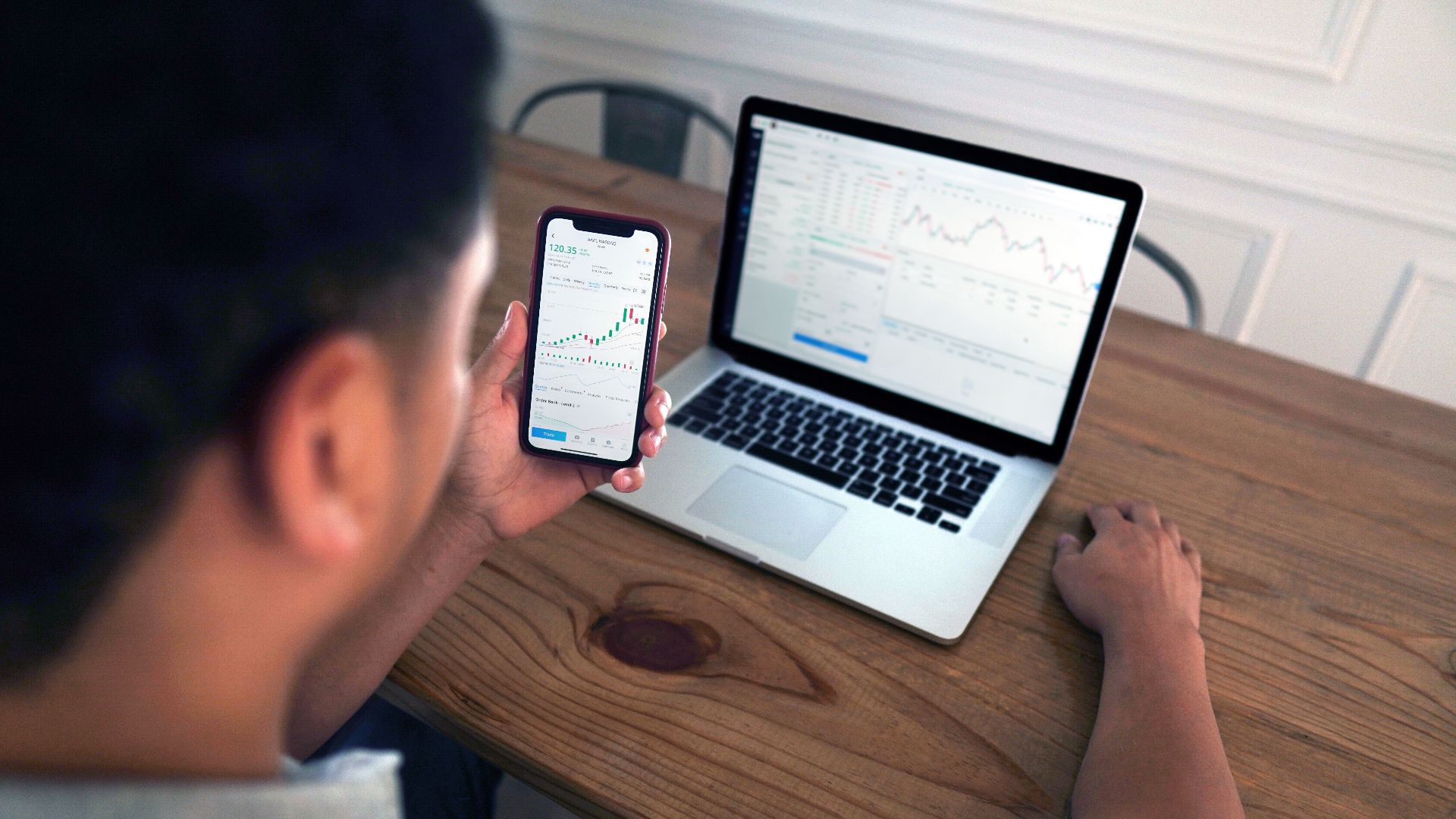 Individual reviewing stock market trends using a laptop and smartphone at a wooden table.