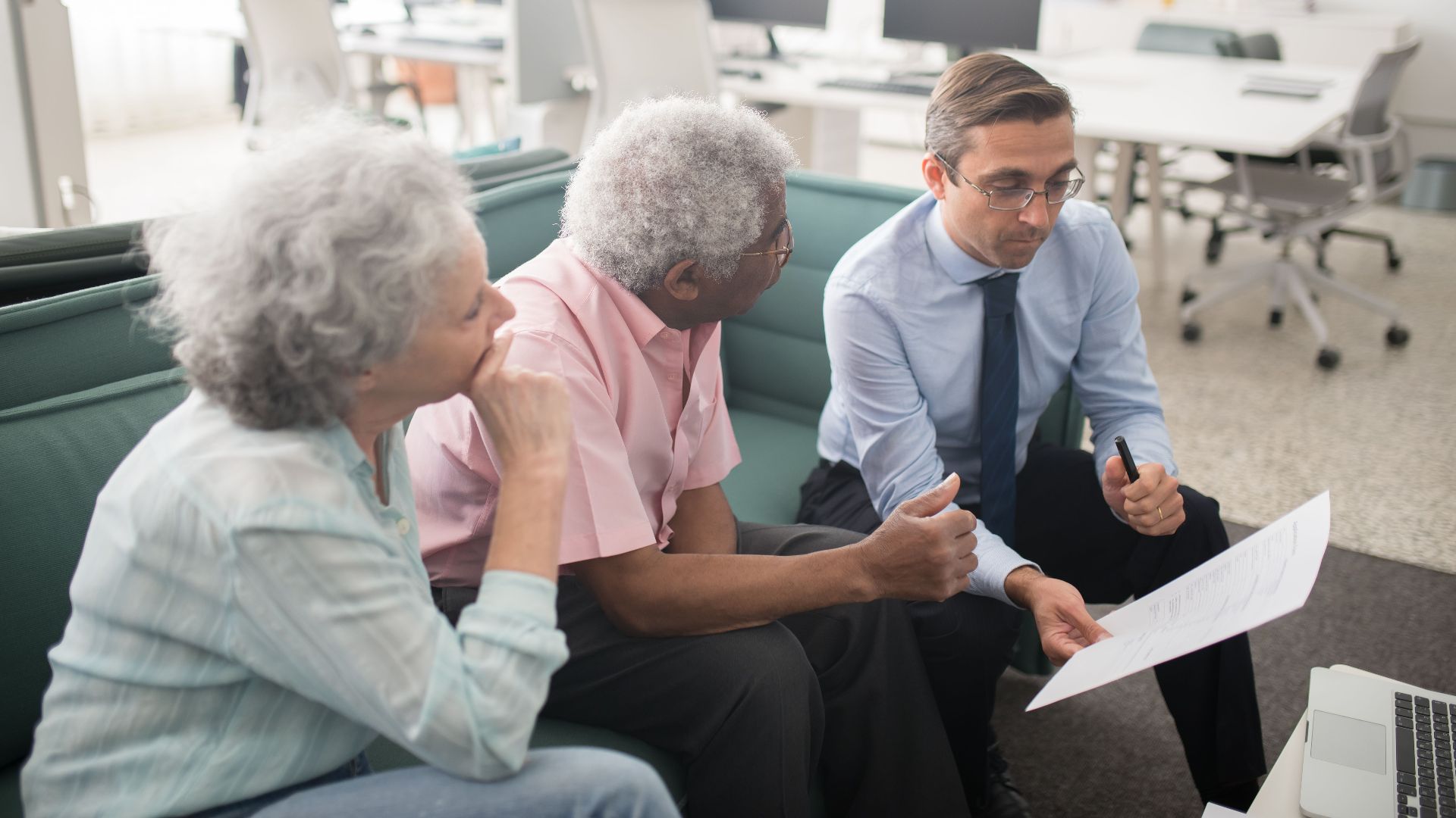 Business professional consults elderly clients in an office setting. Collaborative discussion, paperwork visible.