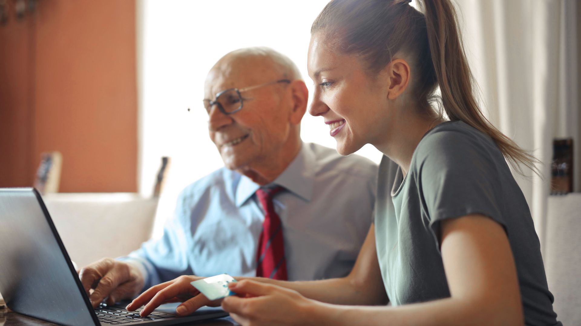 Young woman in casual clothes helping senior man in formal shirt with paying credit card in Internet using laptop while sitting at table