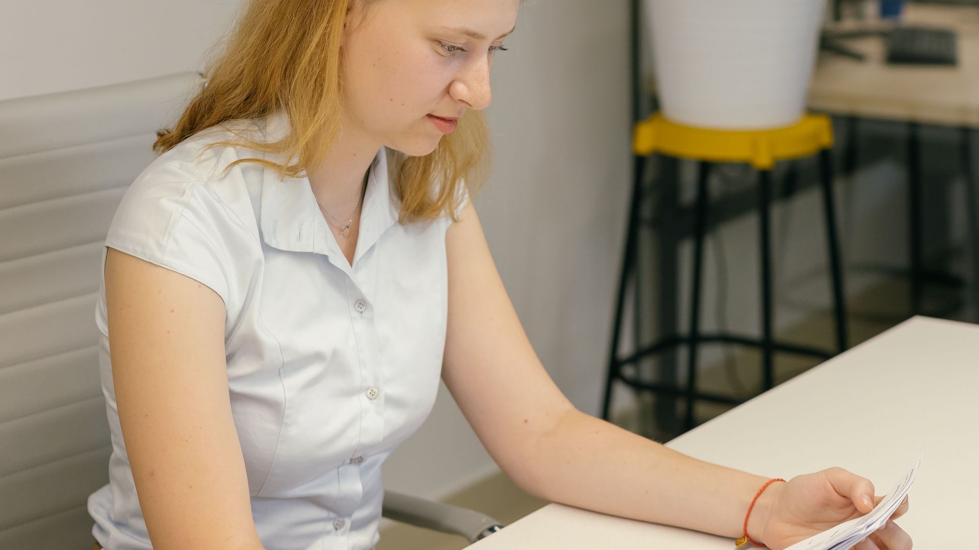 A woman in a modern office reading documents at a desk beside an indoor plant.