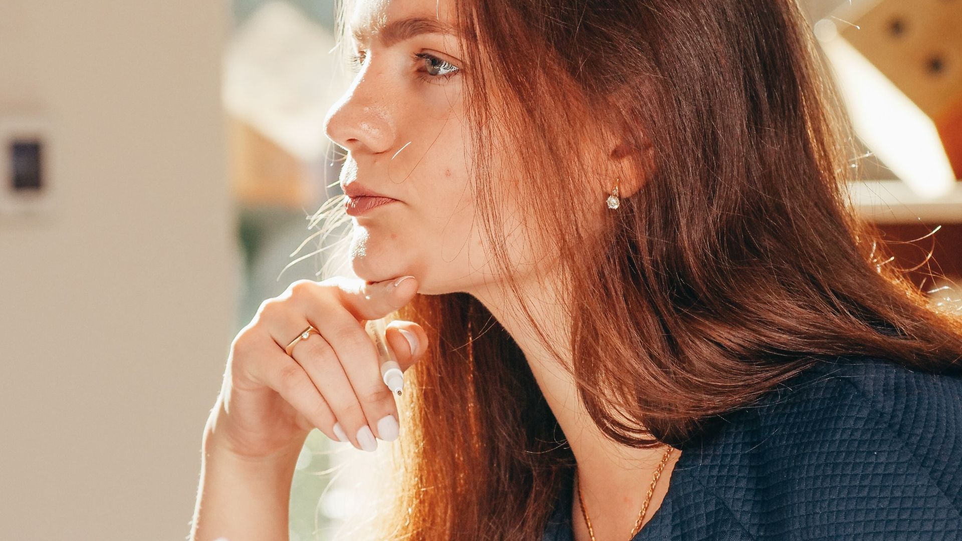 A thoughtful woman looks out the window while holding papers, bathed in warm sunlight indoors.