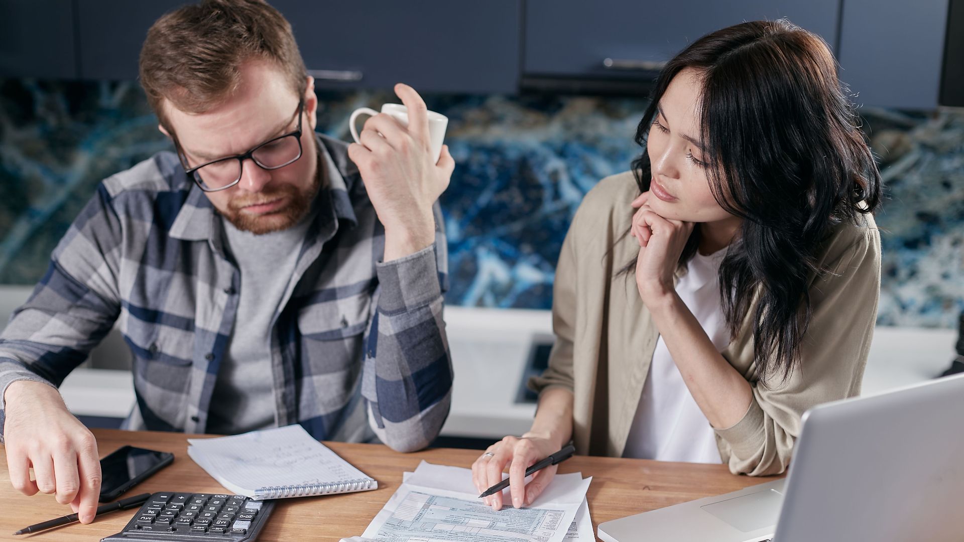 A couple reviews bills and documents on a laptop while discussing household finances.