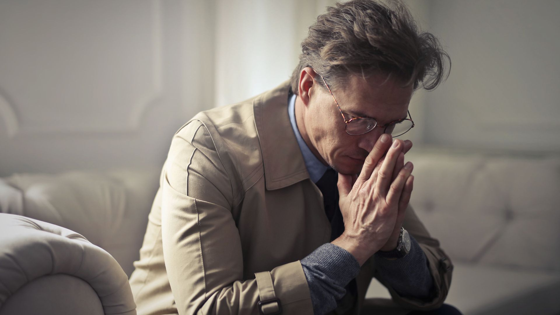 Side view of upset male entrepreneur in formal wear sitting on couch in modern living room and contemplating about financial problems while leaning on hands and looking down
