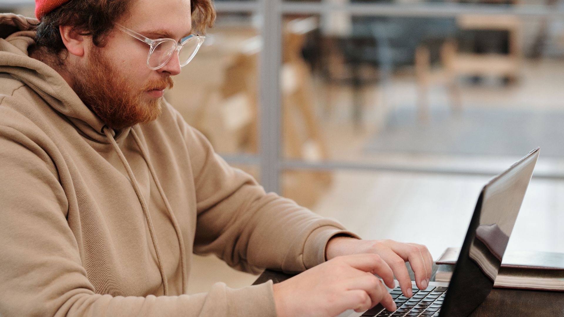 A bearded man wearing glasses, working intently on a laptop in a cafe setting.