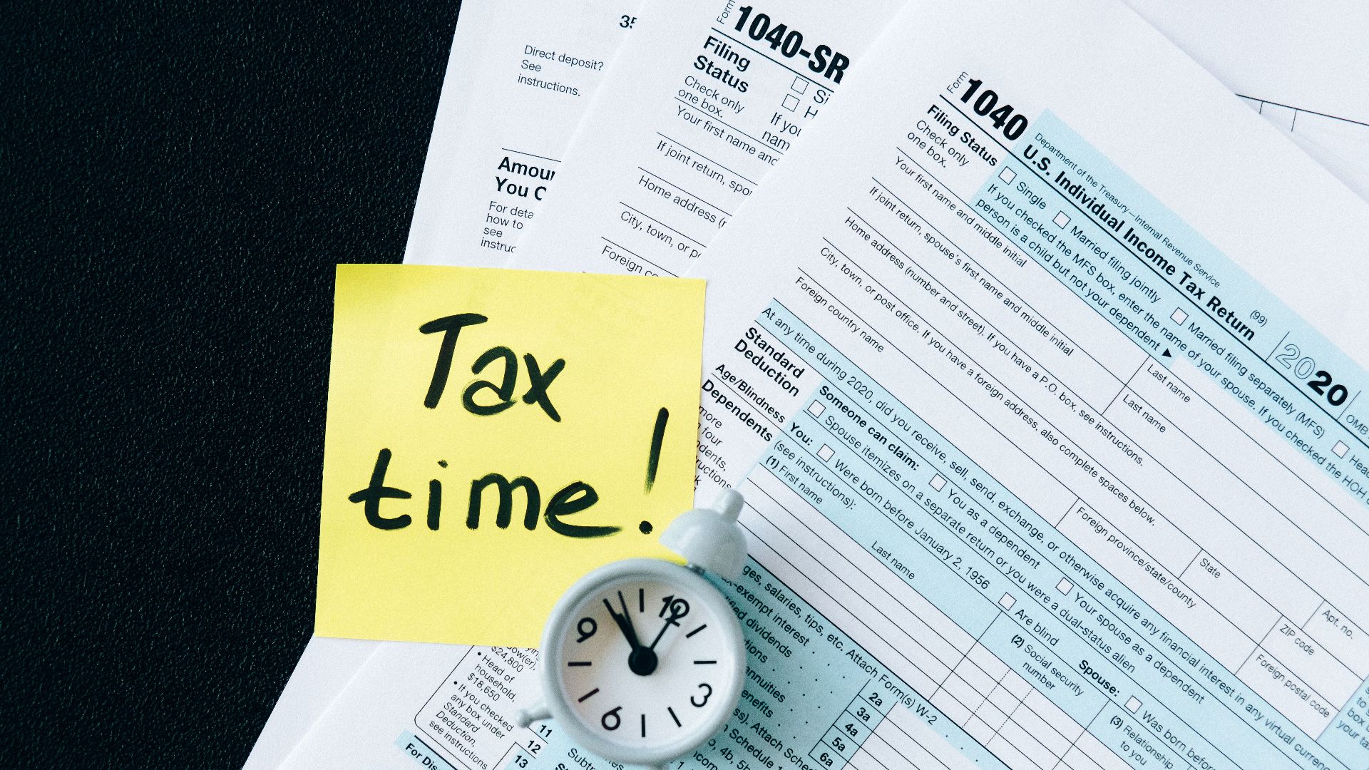 A stack of tax forms with a clock and yellow sticky note saying 'Tax time!' indicating urgency.