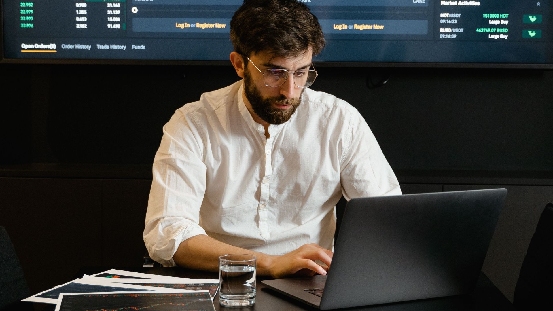 A focused man analyzing stock market trends on a laptop in an office setting with charts displayed.