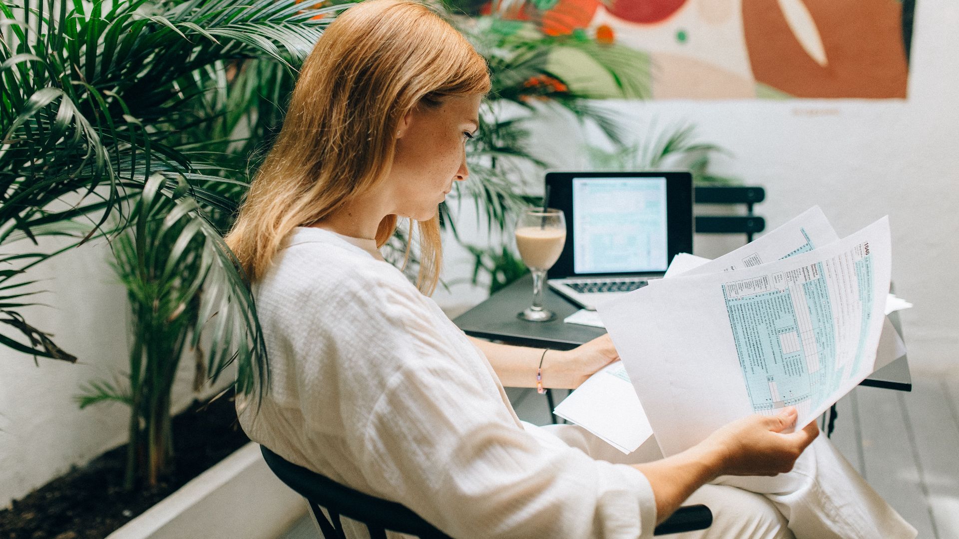 A woman reviews financial documents in an indoor garden space, with a laptop open.