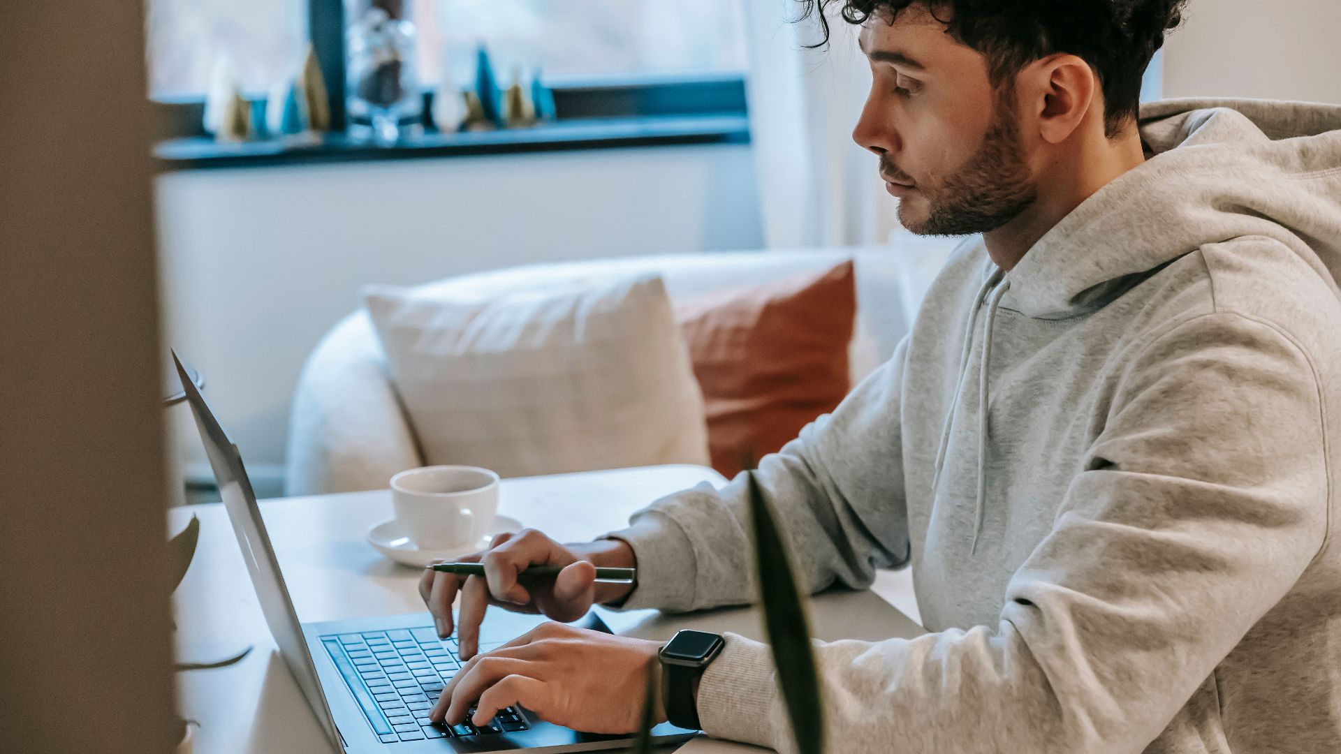 Side view of crop focused male remote worker browsing internet on netbook at table with coffee in house