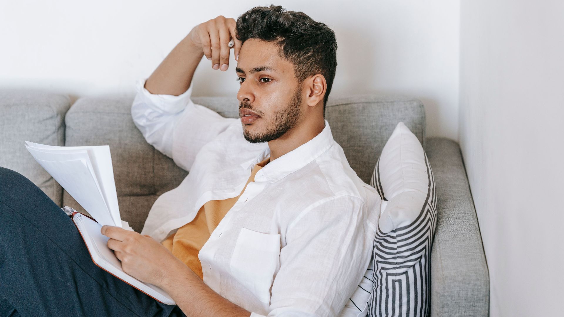 Concentrated young ethnic male wearing casual clothes reading documents and diary and touching head while resting on sofa and looking away