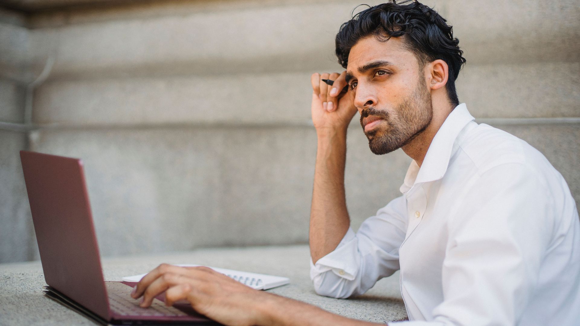 Professional man in a white shirt working on a laptop, contemplating ideas in an urban setting.