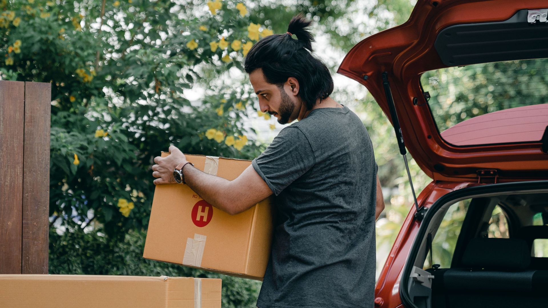 Side view of serious young ethnic man in casual clothes unloading cardboard boxes out of trunk of modern car in blooming green backyard while moving in new house