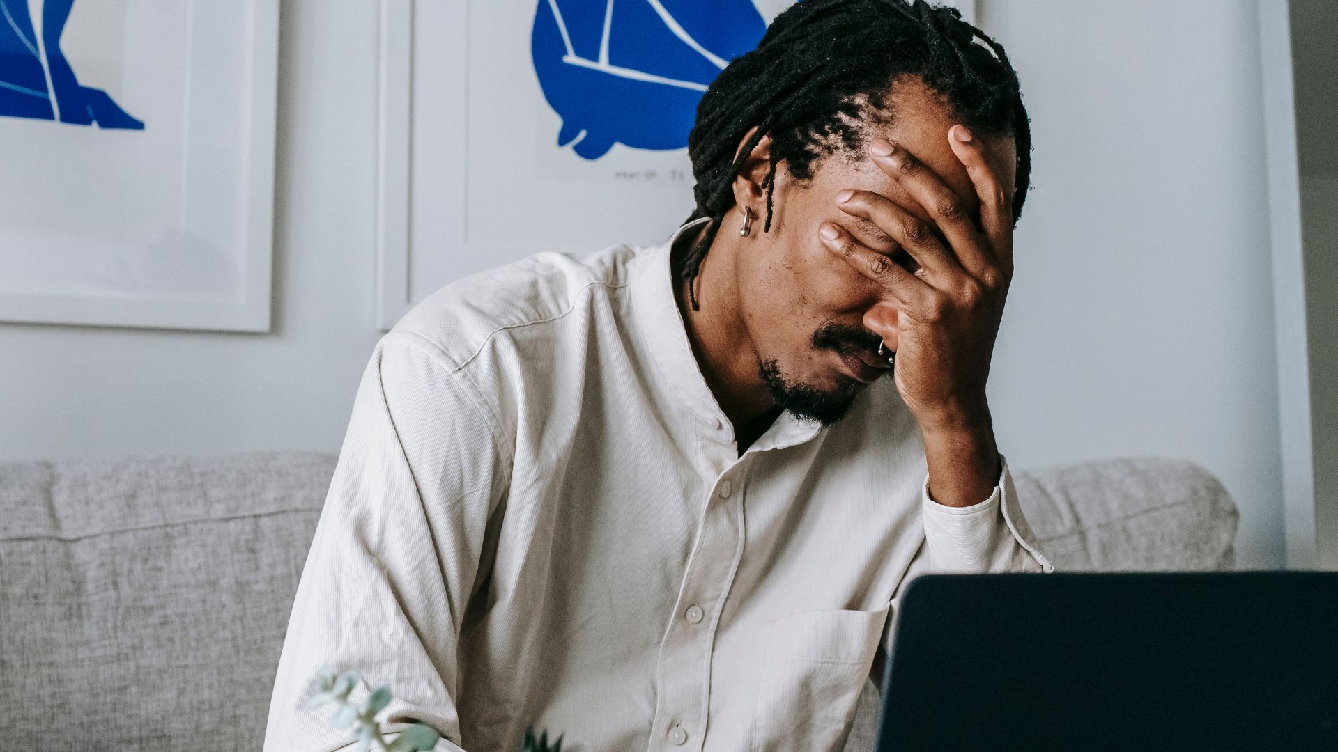 Frustrated young bearded African American male freelancer with dreadlocks in casual shirt working on laptop at home and covering eyes with hand after failure