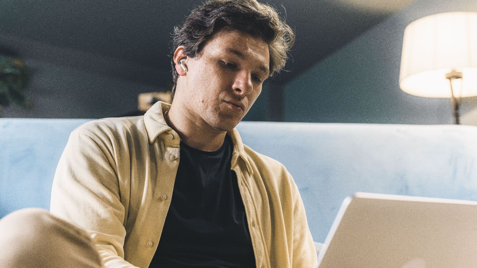A young man sits indoors, working on his laptop with a thoughtful expression, under warm lighting.