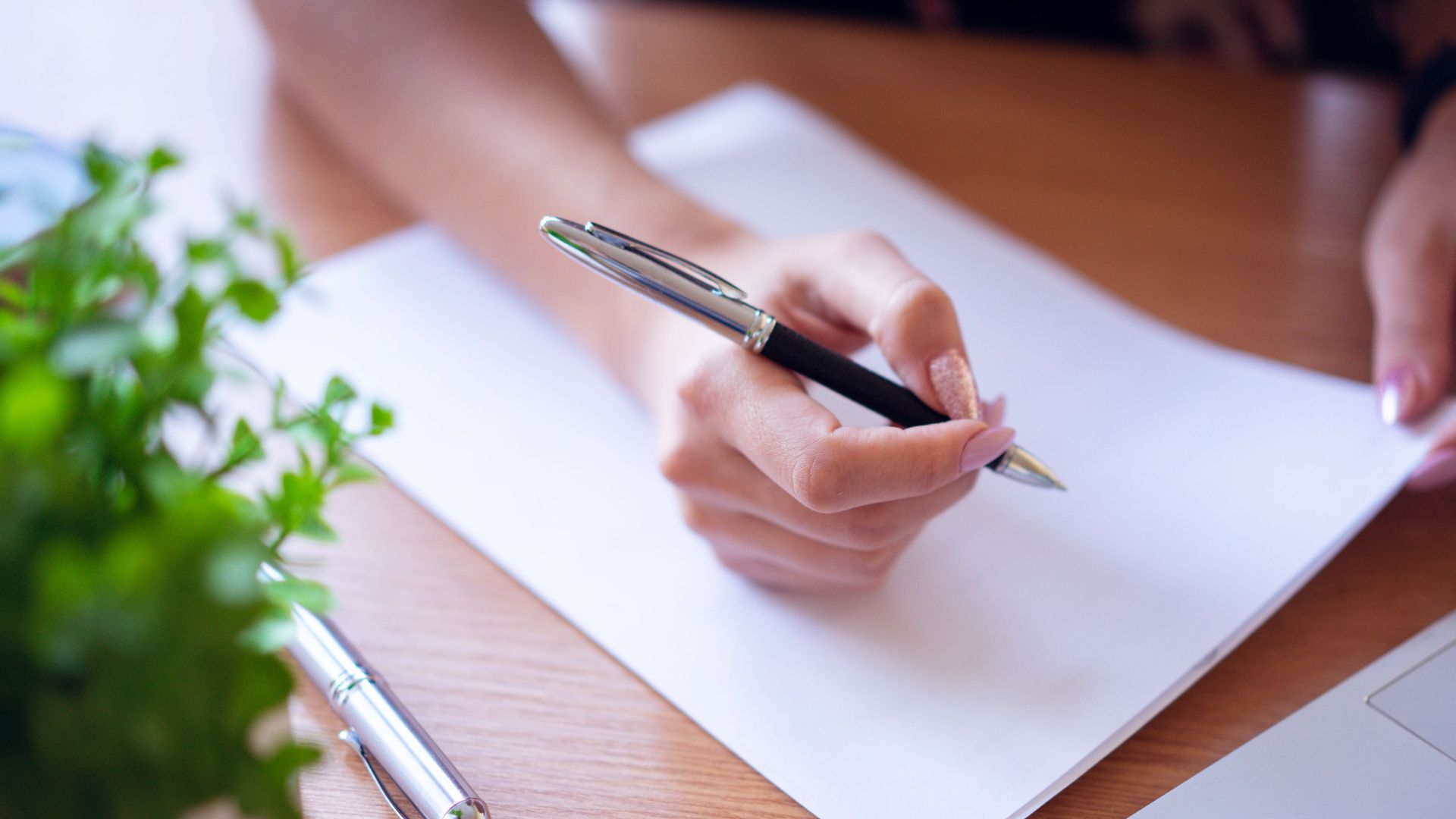 Close-up of a woman writing with a black pen on white paper next to a laptop and plant.