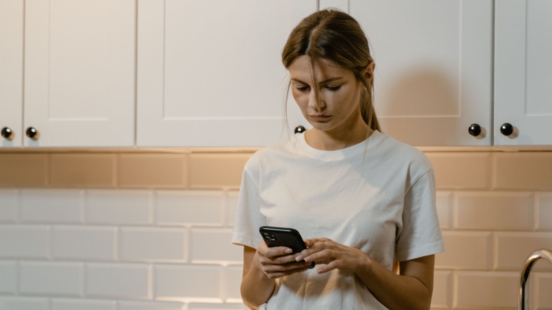 Young woman in kitchen using smartphone, looking focused and thoughtful.