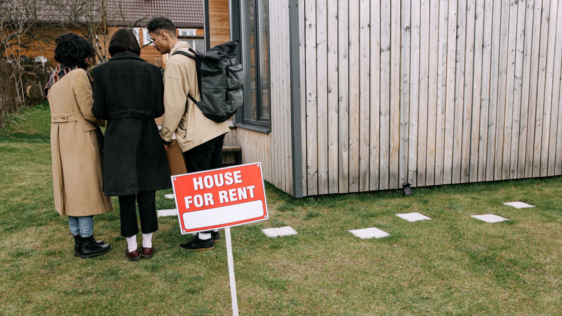 Group of adults viewing a wooden house with a 'House for Rent' sign on the lawn.