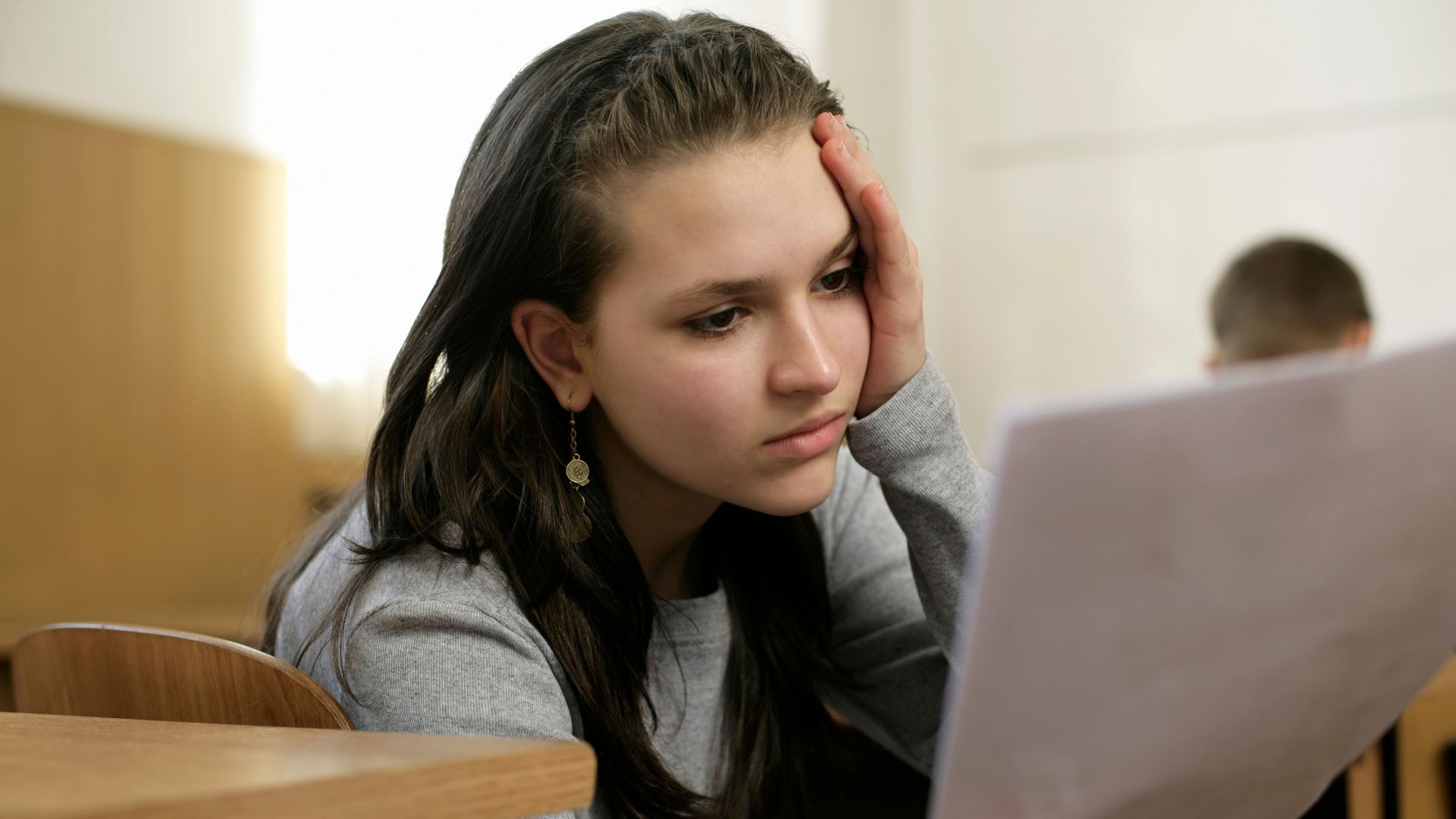 Young woman in a classroom deeply focused on reading a document, holding her head thoughtfully.