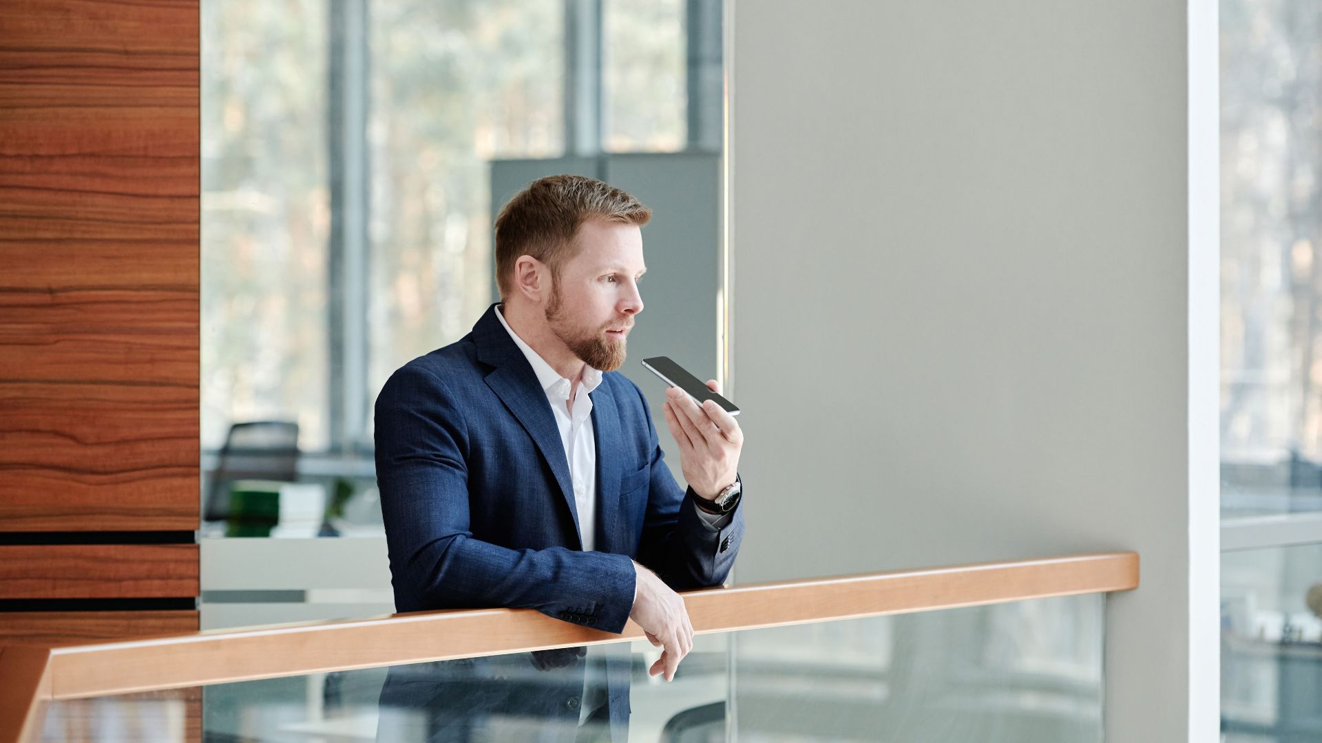 Professional businessman holding a smartphone and speaking indoors at a modern office setting.