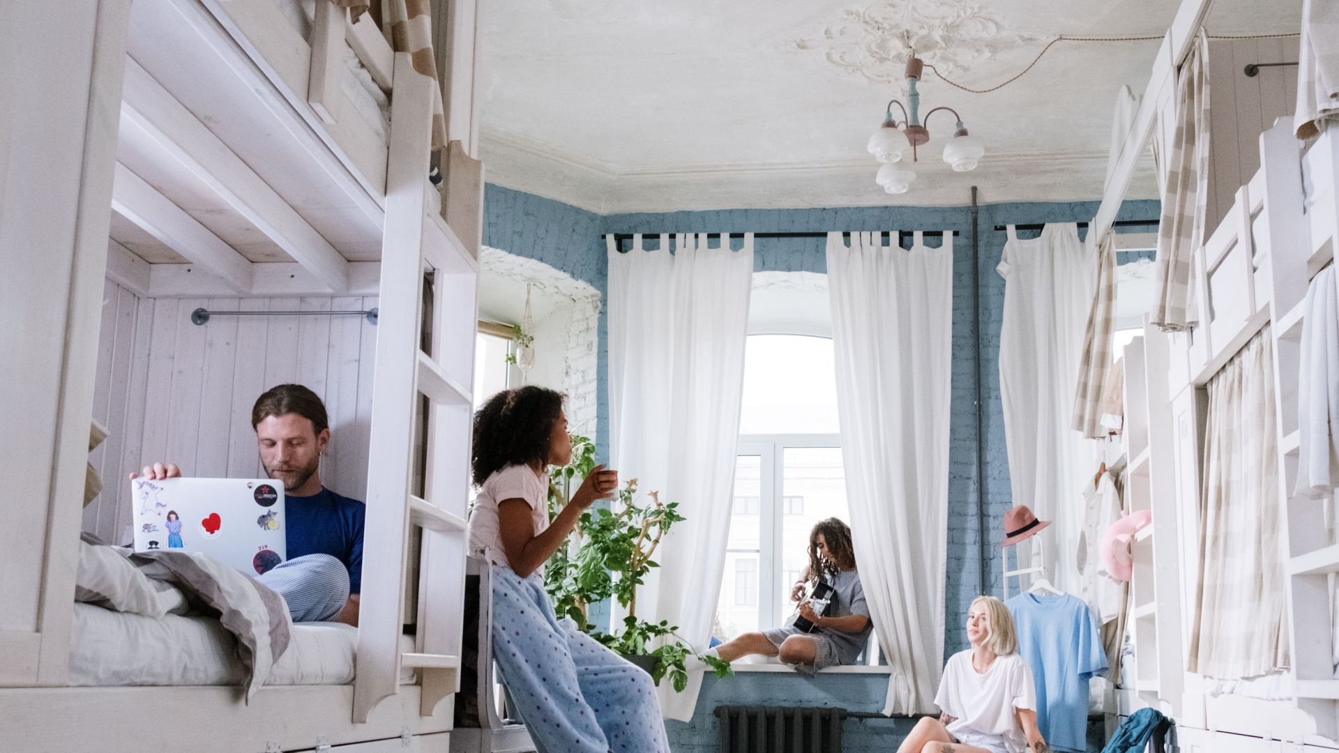 A cozy dorm room where friends relax, chat, play guitar, and use laptops together. A casual hangout environment.