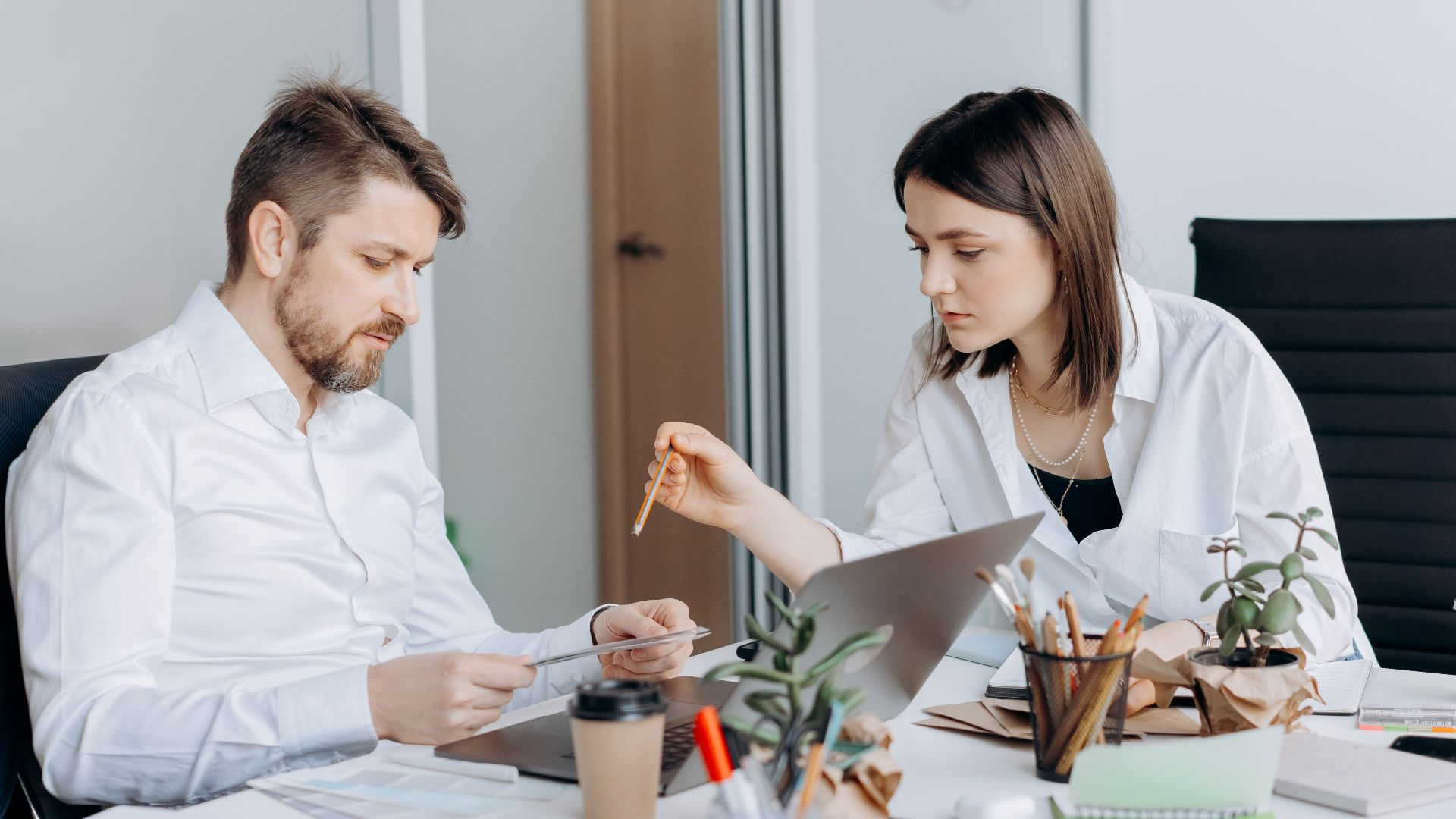 Two colleagues discussing work at a desk with laptops and coffee in a modern office setting.