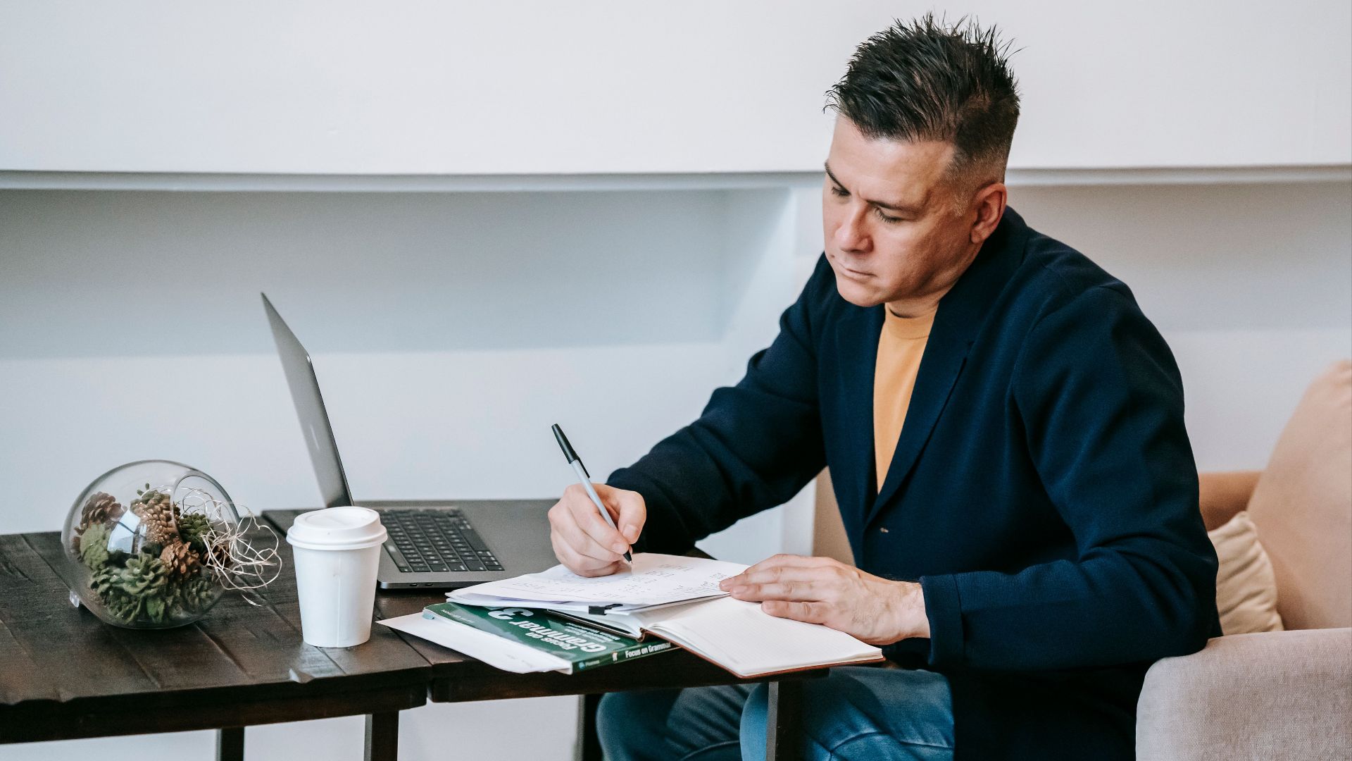Man diligently working in a home office setting, taking notes with a laptop and coffee.