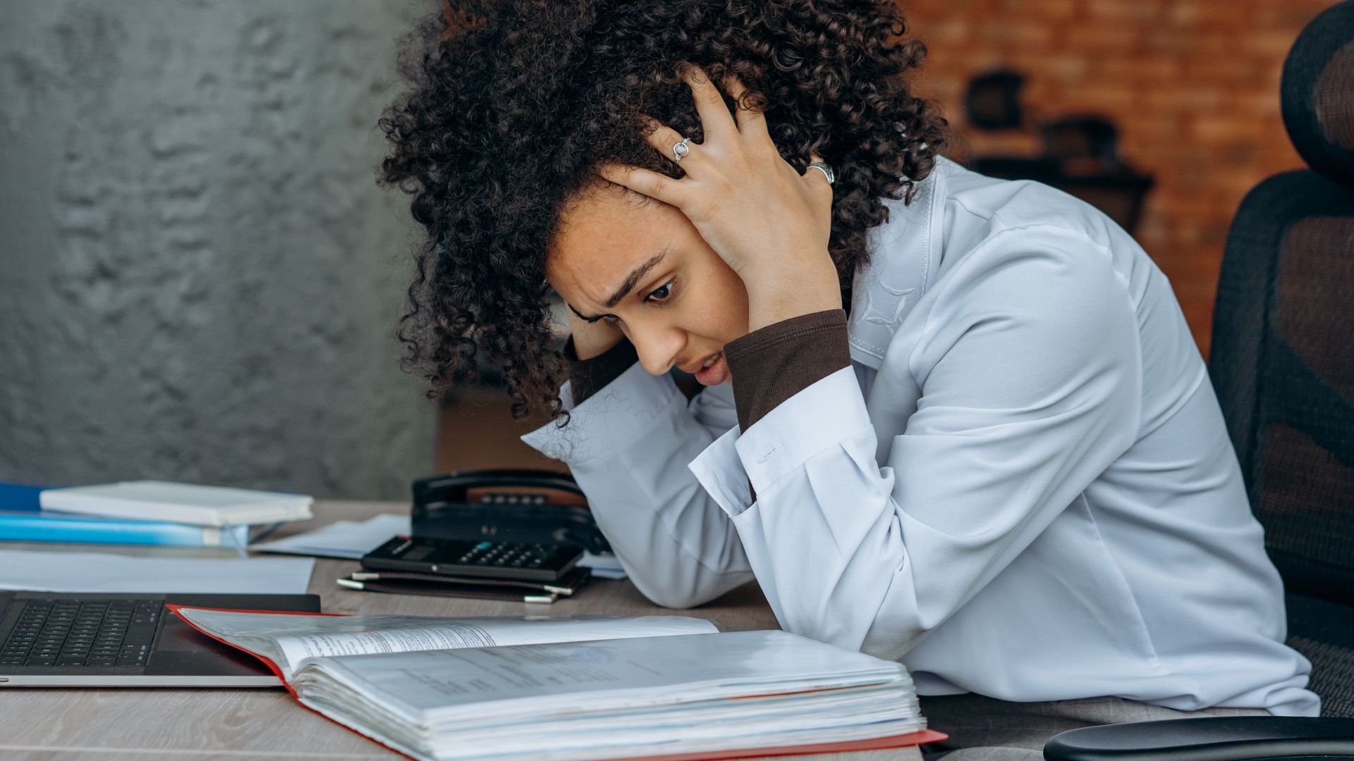 Woman sitting at desk appearing stressed while reviewing documents in an office setting.