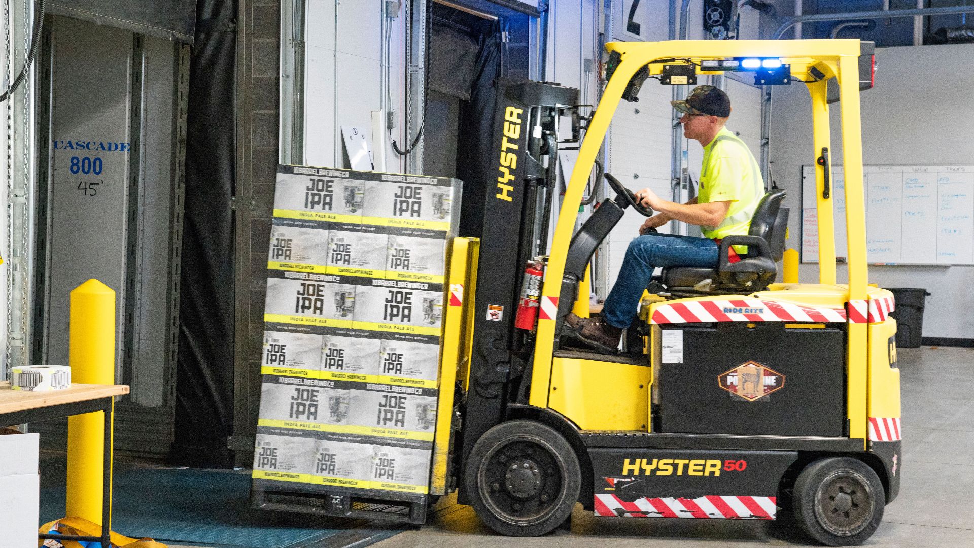 A worker drives a Hyster forklift moving Joe IPA boxes in a warehouse.