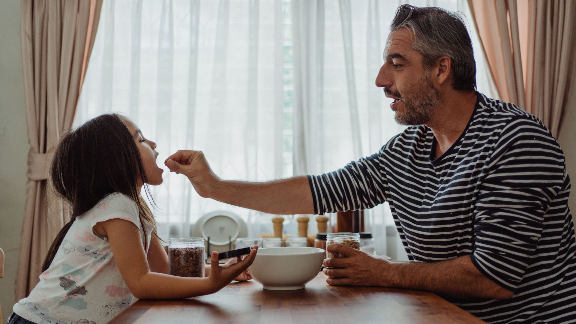 A father playfully feeds his daughter at a dining table, showcasing warmth and family bonding.